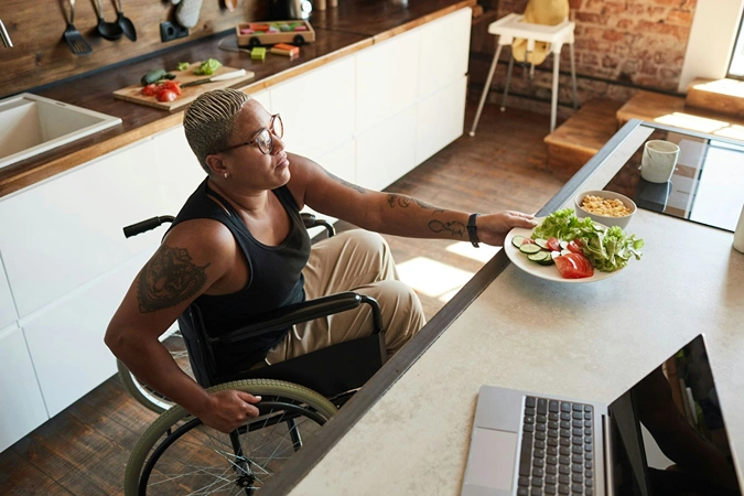 In a kitchen, a woman in a wheelchair is eating a salad, highlighting accessibility and healthy eating.