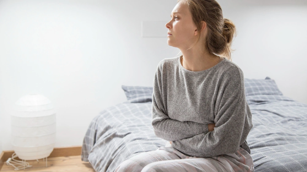 Woman sitting on a bed with stomach discomfort, representing daily life with IBD and Crohn’s disease.